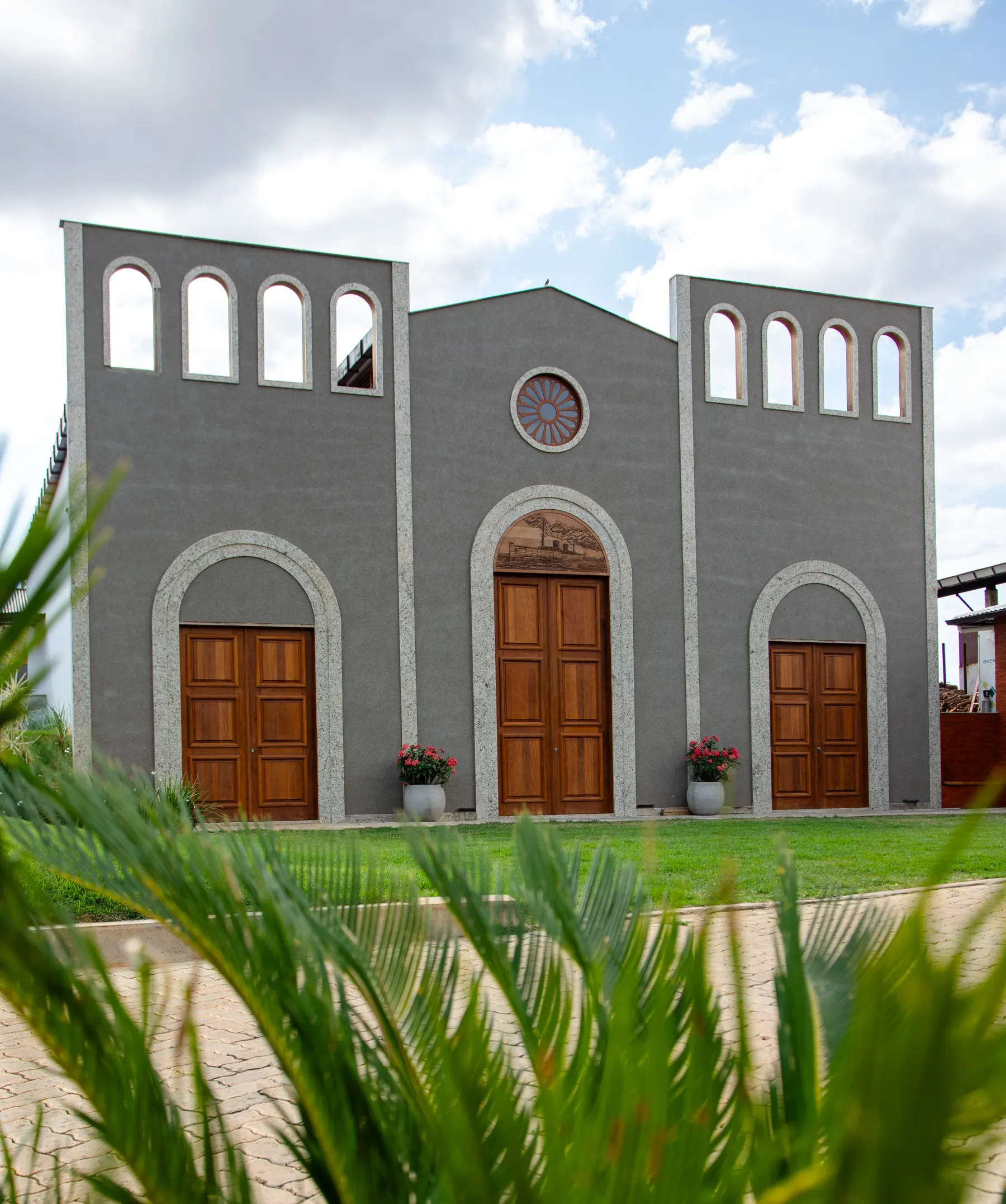 Fachada cinza com três portas de madeira em arco, rosácea e gramado em frente